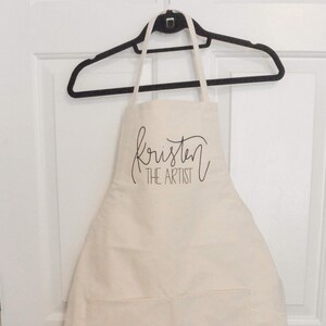 May include: A white cotton apron with the text "Kristen the Artist" in black cursive lettering.