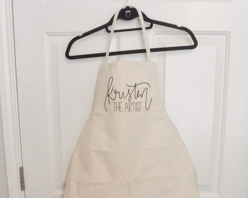 May include: A white cotton apron with the text "Kristen the Artist" in black cursive lettering.