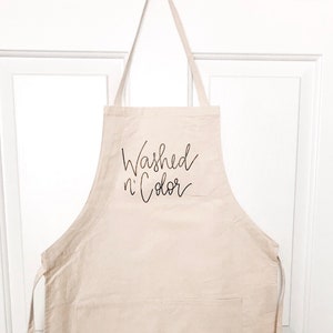 May include: A beige canvas apron with a large pocket and adjustable straps. The apron has the text "Washed n' Color" printed in black on the front.