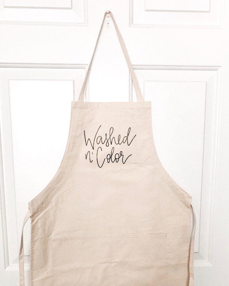 May include: A beige canvas apron with a large pocket and adjustable straps. The apron has the text "Washed n' Color" printed in black on the front.