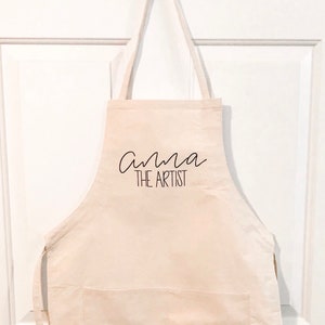 May include: A white canvas apron with a pocket and adjustable straps. The text "Anna the Artist" is printed in black on the front.