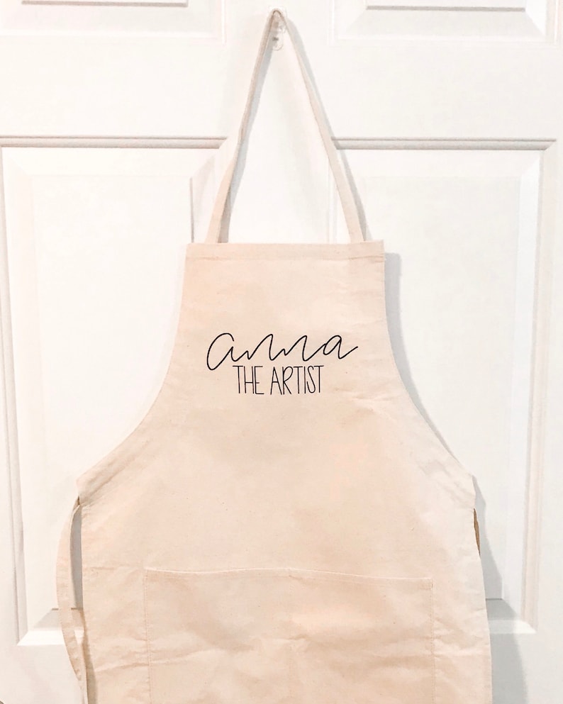 May include: A white canvas apron with a pocket and adjustable straps. The text "Anna the Artist" is printed in black on the front.