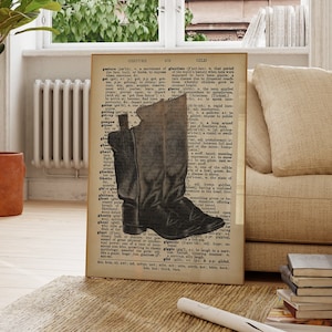 May include: A framed print featuring a black and white illustration of a cowboy boot overlaid on a vintage dictionary page. The artwork is displayed in a well-lit room with a plant and a neutral-toned rug.