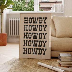 May include: A framed print with the word "HOWDY" repeated five times in black text on a beige background. The print is hanging on a white wall in a living room setting with a couch, a plant, and a chair.