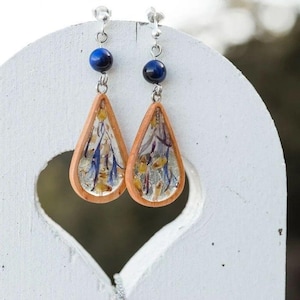 Op de afbeelding: Een paar handgemaakte oorbellen. Elke oorbel heeft een druppelvormig houten frame gevuld met gedroogde bloemen in blauw, geel en paars. Een zilveren ketting verbindt het houten frame met een donkerblauwe kraal en een zilveren pin.