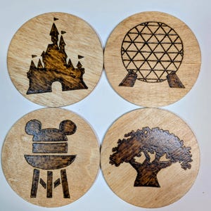May include: Four round wooden coasters with dark brown etched designs. One features a castle, another a geodesic sphere, a third a Mickey Mouse hat on a chair, and the last a tree. The coasters are made of light-colored wood.