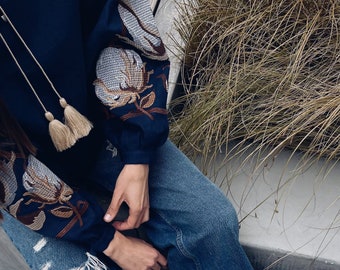 May include: A navy blue embroidered blouse with long sleeves and a tassel detail. The blouse is paired with blue jeans and white sneakers.