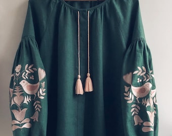 May include: A dark green blouse with long, puffed sleeves and detailed embroidery on the sleeves. The embroidery showcases birds and floral patterns in light pink. The blouse has a round neckline and a drawstring with tassels.