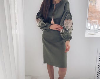 以下が含まれることがあります： 女性が、袖に刺繍された花柄のディテールが施された、オリーブグリーンのロングスリーブドレスを着ています。ドレスは丸首で、前面にタッセルが付いています。