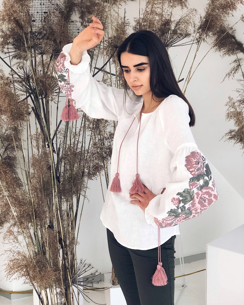 Op de afbeelding: Witte blouse met lange mouwen en bloemenborduurwerk op de mouwen. De blouse heeft roze kwastjes en een ronde halslijn. De mouwen hebben een gerimpelde manchet. De bloemenborduurwerk bevat roze, groene en grijze tinten.