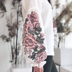 Op de afbeelding: Witte linnen blouse met lange mouwen met geborduurde bloemmotieven in roze, bordeaux en groen. De mouwen hebben een geplooid ruchedetail bij de pols. De blouse is een klassieke stijl.