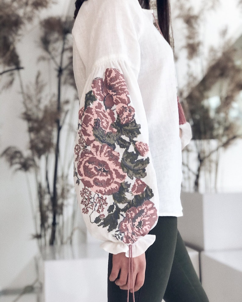 Op de afbeelding: Witte linnen blouse met lange mouwen met geborduurde bloemmotieven in roze, bordeaux en groen. De mouwen hebben een geplooid ruchedetail bij de pols. De blouse is een klassieke stijl.