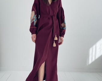 May include: A long, burgundy wrap dress with embroidered floral details on the sleeves. The dress has a tie belt and a side slit. The model is wearing clear heeled sandals.
