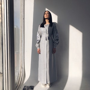 May include: A woman wearing a light blue linen dress with embroidered details and long sleeves. The dress has a button-up front and a tassel detail at the neckline. She is standing in a room with a window and white walls.