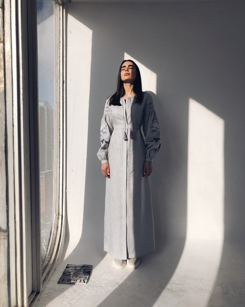 May include: A woman wearing a light blue linen dress with embroidered details and long sleeves. The dress has a button-up front and a tassel detail at the neckline. She is standing in a room with a window and white walls.