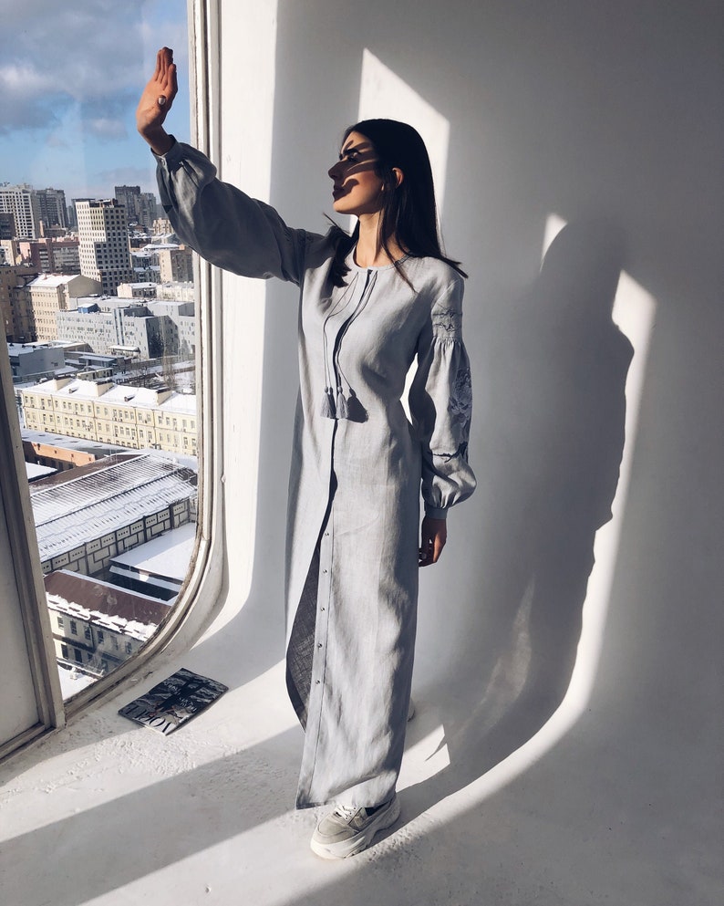 May include: A woman wearing a light blue linen dress with embroidery details and white trainers. She is standing in front of a window with a city view.