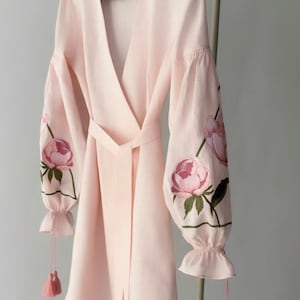 May include: A light pink robe with long sleeves and a wrap-around design. The sleeves are adorned with embroidered pink peony flowers and green leaves. The robe has a matching belt and pink tassels hanging from the sleeves and hem.