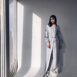 May include: A light blue maxi dress with long sleeves and embroidery on the sleeves. The dress has a button-up front and a slit in the skirt. The model is wearing white trainers.