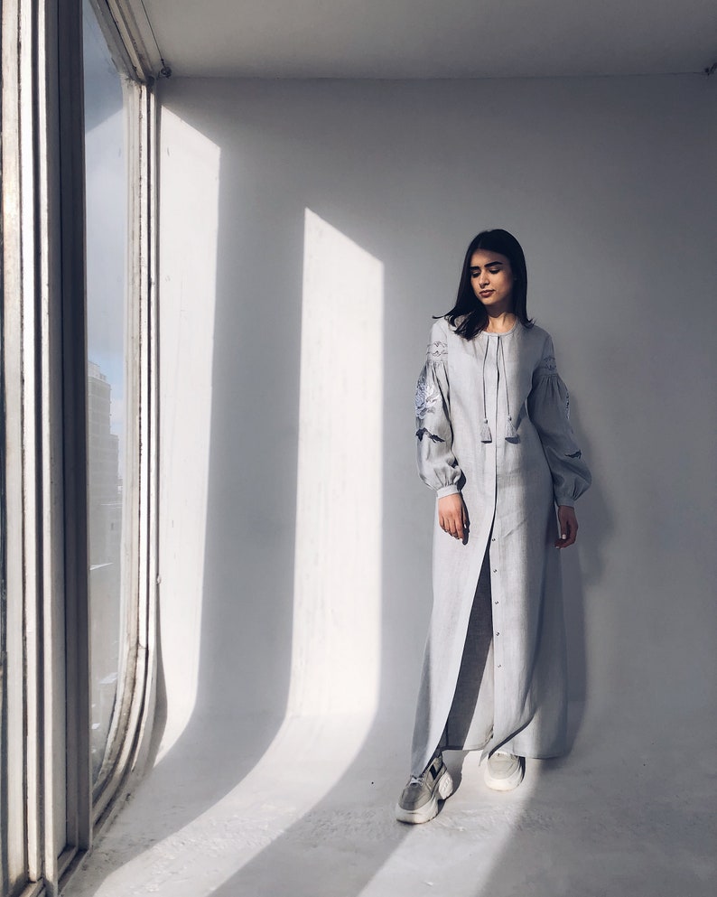 May include: A light blue maxi dress with long sleeves and embroidery on the sleeves. The dress has a button-up front and a slit in the skirt. The model is wearing white trainers.