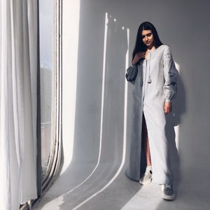 May include: A woman wearing a long, light blue linen dress with embroidered details. The dress has a button-up front and a tassel detail at the neckline. She is standing in a room with a window and white walls.