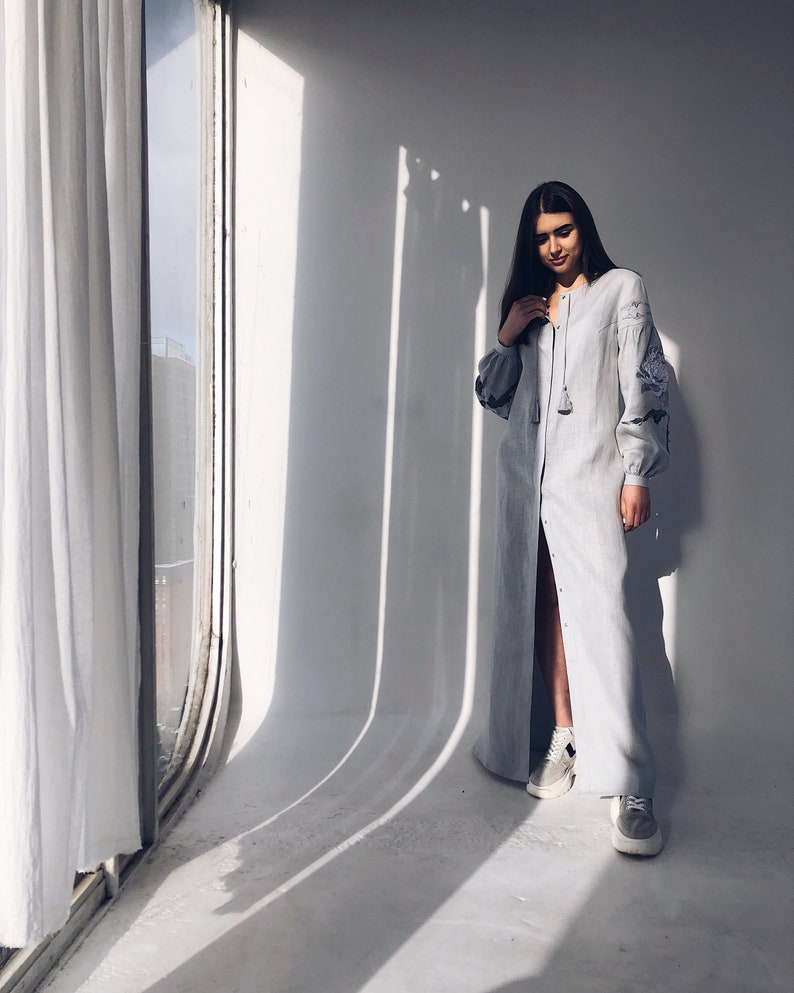 May include: A woman wearing a long, light blue linen dress with embroidered details. The dress has a button-up front and a tassel detail at the neckline. She is standing in a room with a window and white walls.