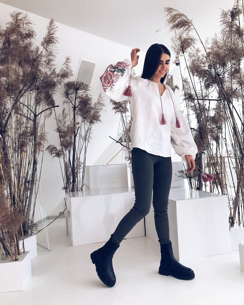 Op de afbeelding: Witte linnen blouse met geborduurde bloemen op de mouwen en kwastjes. Gedragen met een donkergroene legging en zwarte enkellaarsjes. De achtergrond toont hoge, gedroogde planten en een wit interieur.