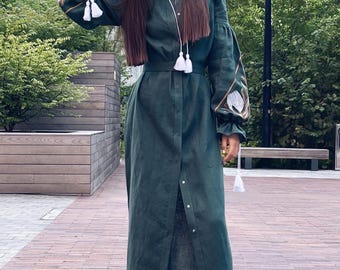 May include: A long, dark green linen dress with white embroidery on the sleeves and a white tassel at the waist. The dress has a button-up front and a relaxed fit. The model is wearing white sneakers.
