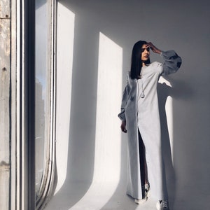 May include: A woman wearing a light blue long-sleeved dress with a slit up the front. She is standing in a room with a large window behind her.