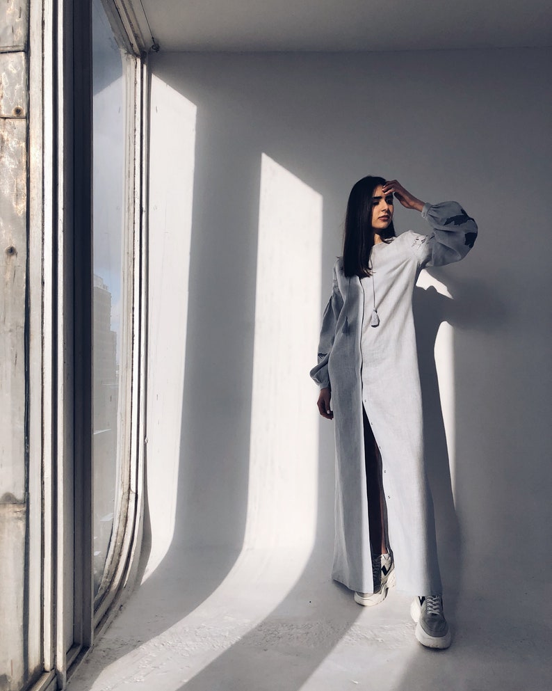 May include: A woman wearing a light blue long-sleeved dress with a slit up the front. She is standing in a room with a large window behind her.