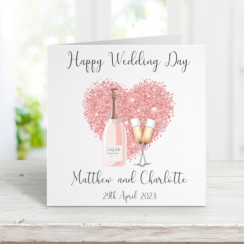 May include: A pink glitter heart with a bottle of champagne and two champagne flutes. The text "Happy Wedding Day" is at the top of the card. The text "Matthew and Charlotte" is below the heart. The date "29th April 2023" is below the names.