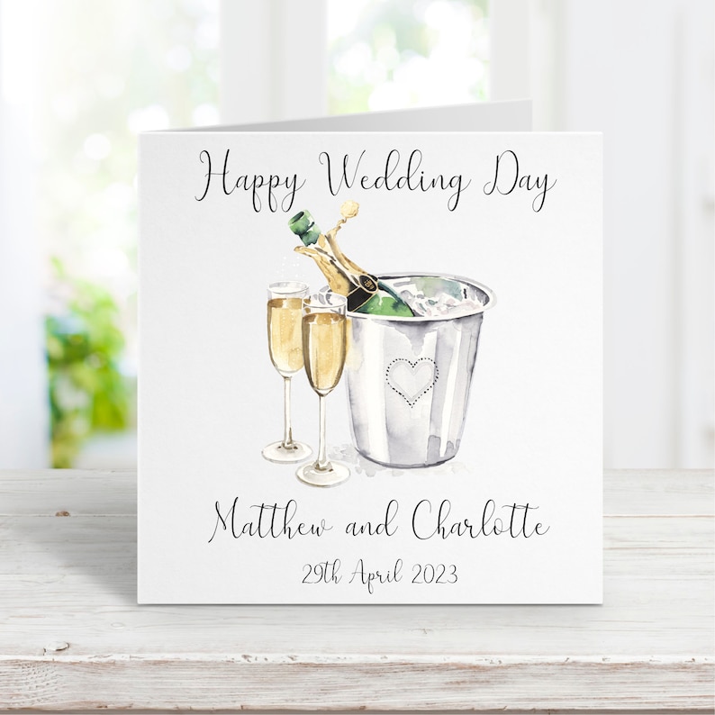 Puede incluir: Una tarjeta blanca con una ilustraci&oacute;n de acuarela de una botella de champ&aacute;n en un cubo de hielo con dos copas de champ&aacute;n. La tarjeta dice "Happy Wedding Day" en la parte superior, "Matthew and Charlotte" en el medio y "29th April 2023" en la parte inferior.