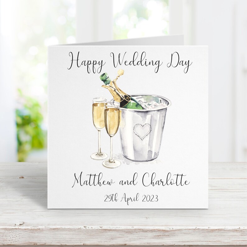 Puede incluir: Una tarjeta blanca con una ilustraci&oacute;n de acuarela de una botella de champ&aacute;n en un cubo de hielo con dos copas de champ&aacute;n. La tarjeta dice "Happy Wedding Day" en la parte superior, "Matthew and Charlotte" en el medio y "29th April 2023" en la parte inferior.