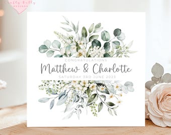 May include: A white card with a watercolor illustration of a bouquet of white flowers and green leaves. The text "Congratulations Matthew & Charlotte Saturday 3rd June 2023" is printed on the card.