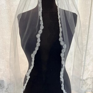 Peut inclure: Un voile de mariée blanc avec une délicate bordure en dentelle florale. Le voile est présenté sur un mannequin noir, mettant en valeur son design fluide. La bordure en dentelle ajoute une touche d'élégance.