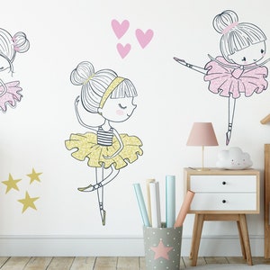 May include: Three ballerina girl wall decals in pink, yellow, and gold glitter. The decals are on a white wall with a pink dresser, a wooden bed frame, and a pink flamingo plush toy.