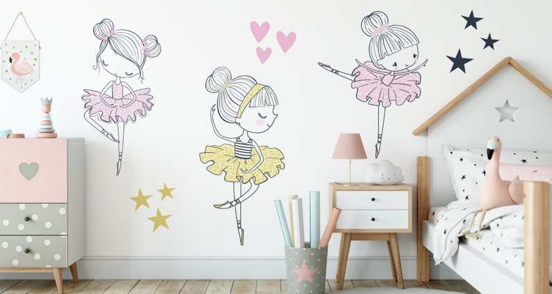 May include: Three ballerina girl wall decals in pink, yellow, and gold glitter. The decals are on a white wall with a pink dresser, a wooden bed frame, and a pink flamingo plush toy.