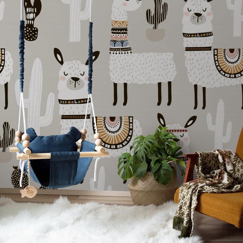 May include: A nursery room with a gray wall featuring a llama pattern. A wooden baby swing with a blue seat hangs in front of the wall. A potted plant sits on the floor next to a brown chair with a green and white throw blanket.
