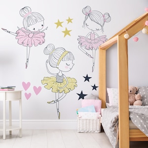 May include: Three ballerina girl wall decals in pink, yellow, and black with gold and black stars. The decals are on a white wall in a bedroom with a wooden bed and a white side table.