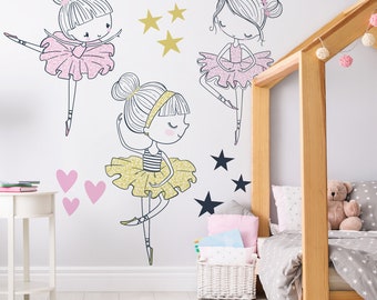 May include: Three ballerina girl wall decals in pink, yellow, and black with gold and black stars. The decals are on a white wall in a bedroom with a wooden bed and a white side table.