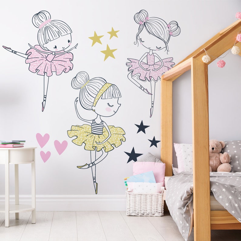 May include: Three ballerina girl wall decals in pink, yellow, and black with gold and black stars. The decals are on a white wall in a bedroom with a wooden bed and a white side table.