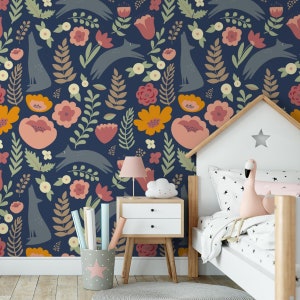 May include: A blue and white wallpaper with a repeating pattern of stylized foxes, flowers, and leaves. The wallpaper is shown in a child's bedroom with a white bed, a nightstand, and a pink flamingo toy.