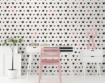 May include: A modern home office setup with a white desk, chair, and side table. The wall is covered in a white wallpaper pattern with black and pink hearts. A pink lamp and a potted plant add color to the room. The desk has a few books and a small organizer.