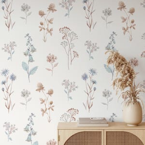 K&ouml;nnte beinhalten: Eine wei&szlig;e Wand mit einem sich wiederholenden Muster aus Wildblumen im Aquarellstil in Braun-, Blau- und Rosat&ouml;nen. Vor der Wand steht ein Holzschrank mit einer Vase mit getrockneten Pflanzen und B&uuml;chern.