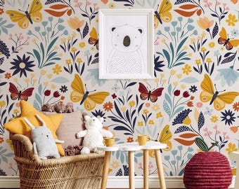 May include: A children's room with a white wall covered in a colorful floral wallpaper featuring butterflies, flowers, and leaves. The wallpaper has a whimsical and playful design. There is a wicker chair with pillows, a small table, and a red apple-shaped basket.