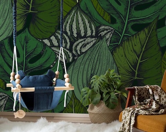 May include: A wooden baby swing with a blue fabric seat hangs in front of a green and white tropical leaf patterned wall. A potted plant and a chair with a throw blanket are in the foreground.