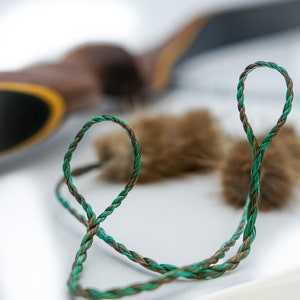 Pode incluir: Close-up de um cordão trançado em tons de verde e castanho, formando um laço. O cordão está em foco, contrastando com um fundo desfocado com um objeto castanho e pequenos elementos castanhos difusos. A iluminação realça a textura.