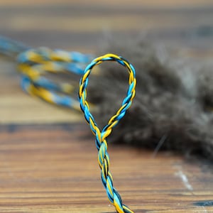 May include: Close-up of a braided cord in a loop shape. The cord is made of strands in yellow, blue, and black. The background is a blurred wooden surface and a soft, gray, feathery texture.