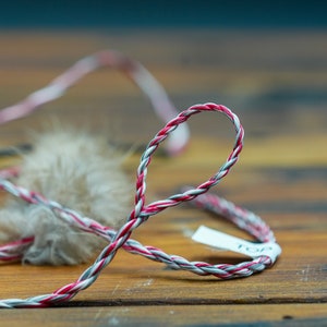 May include: Close-up of a braided cord with red, white, and gray strands, forming a loop. A small, fluffy, tan object is attached to the cord. A white tag with the word "TOP" is attached to the cord. The cord is on a wooden surface.