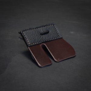 Può includere: Primo piano di un paracordino in pelle per il tiro con l'arco. Il paracordino presenta una parte superiore in pelle blu scuro con un bordo cucito e un proteggi dita in pelle marrone. Un cordino nero è attaccato alla parte superiore. Sfondo scuro.
