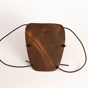 Könnte beinhalten: Ein brauner Lederarmschutz mit schwarzen Kordelbändern. Der Schutz hat eine gebogene Form und eine strukturierte Oberfläche. Das Leder hat eine warme braune Farbe mit dunkleren Variationen. Der Text "JAMES HENDERSON" ist auf dem Leder geprägt.